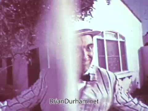 The still from the 1968 Red Rose Tea commercial features a man in a striped outfit, smiling as he engages with the camera. The image is processed in black and white but originally in color, offering a nostalgic, lively atmosphere typical of that era. The background suggests a residential setting, with a hint of greenery and a house. The composition captures a playful moment, highlighting the charm of vintage advertising.