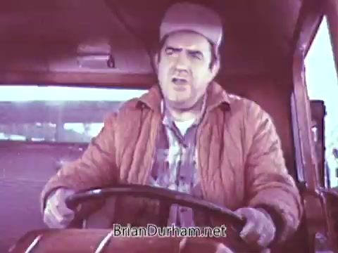 The still depicts a man driving a vehicle, likely in a commercial setting. He is wearing a cap and a quilted jacket, appearing engaged or animated in conversation. The color processing suggests a warm, nostalgic look characteristic of the late 1960s. The environment appears to have a vintage aesthetic, typical of promotional content from that era.