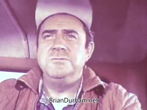 The still from the 'Reupload - Red Rose Tea commercial 1968' features a close-up of a man in a vehicle, likely a truck. He wears a light-colored cap and a textured jacket, conveying a serious expression. The background suggests an outdoor setting, although details are muted due to the black-and-white processing. The overall mood appears contemplative, reflecting the tone often found in commercials of that era.
