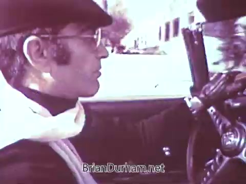 The still features a man driving a convertible car, dressed in a stylish black outfit complemented by a white scarf. He wears sunglasses and a hat, adding a touch of sophistication. The background shows a sunny street, suggesting a leisurely drive. The overall aesthetic reflects mid-20th-century fashion and style.