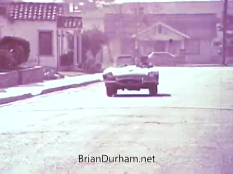 The still shows a vintage convertible car driving down a quiet street, likely in a suburban setting. The scene has a nostalgic feel, with a pale color palette that suggests a sunny day. In the background, there are modest houses and trees lining the street, contributing to the calm and inviting atmosphere typical of the 1960s. The car is the focal point, evoking a sense of leisure and classic Americana.