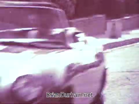 The still from the 'Red Rose Tea' commercial features a vintage car moving on a street. The image showcases a slightly faded look, likely due to the processing in black and white. The scene captures a moment of action, emphasizing the car's presence. Elements in the background hint at a suburban setting, but the overall color vibrancy is lost in this monochrome rendition.