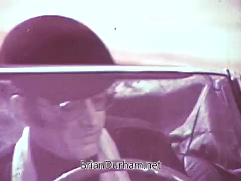 The still from the 1968 Red Rose Tea commercial features a man wearing a bowler hat and sunglasses, seated in a convertible car. The scene captures a bright and colorful atmosphere, likely intended to evoke a sense of enjoyment and leisure associated with the product. The choice to process it in black and white contrasts with the original vibrant colors, altering the intended visual impact.