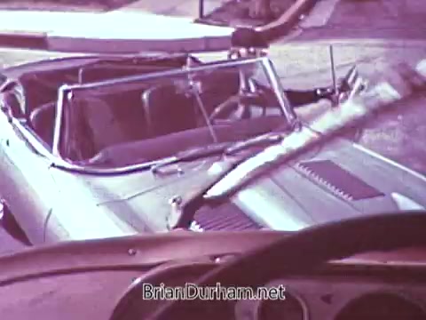 The still shows an open convertible car, possibly set in a sunny outdoor location. The interior appears spacious, with a focus on the front seats. There might be a sense of nostalgia, given the vintage style of the car. The visual quality suggests it's from a 1968 commercial promoting Red Rose Tea, although it is processed in black and white instead of color.