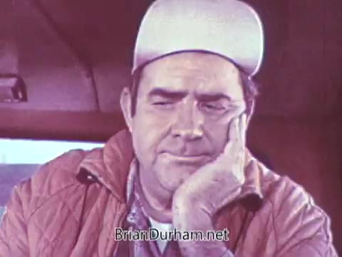 The still features a contemplative man resting his chin on his hand, wearing a light-colored cap and an orange jacket. He appears to be in a vehicle, gazing out with a thoughtful expression, likely reflecting on something significant. The background is slightly blurred, suggesting a focus on his demeanor. This moment captures a sense of nostalgia and introspection, typical of commercials from that era.