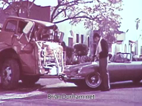 The still from the 1968 Red Rose Tea commercial depicts a man standing next to a light-colored convertible parked on a street. He appears to be inspecting or interacting with the vehicle, which is positioned close to a large, damaged truck. Surrounding the scene are trees and buildings typical of a suburban setting. The image has been processed in black and white, despite originally being colorful.