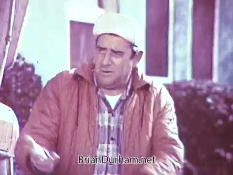 In this still from the 1968 Red Rose Tea commercial, a man is depicted wearing a light-colored cap and an orange jacket over a plaid shirt. His expression appears concerned or contemplative. The background features some greenery, likely shrubs or a garden, giving a glimpse of a warm, outdoor setting. The processing as black and white may have altered the vibrant colors originally present in the scene.
