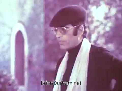 The still appears to feature a man dressed in a black turtleneck and a hat, wearing glasses. He has a scarf draped around his neck, suggesting a stylish, possibly vintage look typical of the late 1960s. The background is likely softly blurred or in muted colors, adding a nostalgic atmosphere to the scene.   The original commercial was meant to showcase Red Rose Tea, highlighting its appeal through vibrant color, which contrasts with the black and white processing here.