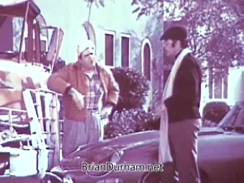 In the 16mm still from the 1968 Red Rose Tea commercial, two men are standing beside a car. One man, dressed casually in a jacket and hat, appears to be talking to the other man, who is wearing a plaid shirt and has a more relaxed demeanor. The background features a building with large windows and some greenery, giving a sense of an outdoor setting. The scene conveys a casual, conversational atmosphere.