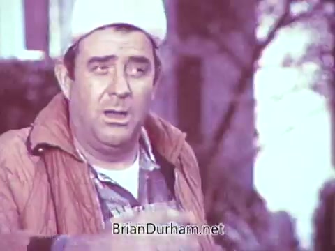 The still from the 1968 Red Rose Tea commercial features a man in a light-colored cap and a quilted jacket, looking animated as he delivers a line. The background suggests a casual outdoor setting, but due to the processing in black and white, the original vibrant colors are lost, creating a monochromatic effect. The scene captures a moment of communication, possibly emphasizing the warmth and character associated with the brand.