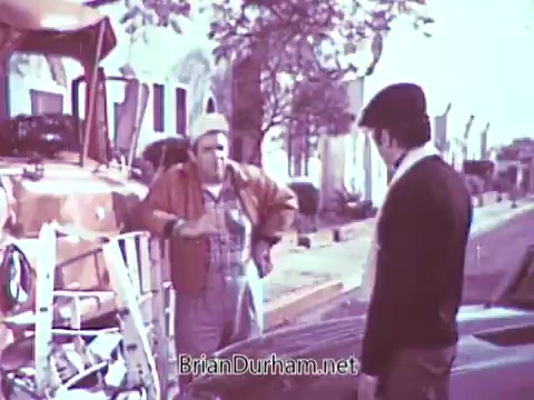 The still depicts two men interacting in a suburban setting. One, dressed in a jacket and hat, appears to be listening intently while the other, in overalls, gestures, possibly explaining something. The scene is set near a truck, with a background of houses and trees. The overall aesthetic, originally in color, has been processed in black and white, altering the visual impact of the lively commercial.