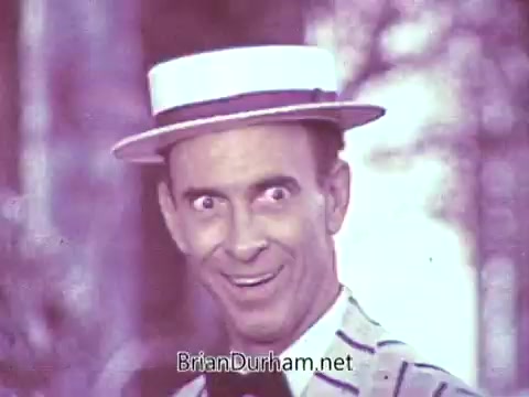The still features a man wearing a straw boater hat and a striped suit, grinning with a playful expression. His eyes are wide and exaggerated, contributing to a lively and humorous tone. The background appears blurred, emphasizing his animated expression. The image, originally in color, has a washed-out quality due to the black-and-white processing.