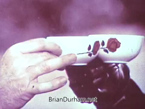 The still shows two hands holding a teacup featuring a rose design. One hand is gloved, contrasting with the bare hand, suggesting an intimate or ceremonial moment. The background is likely blurred, focusing on the cups. The original scene was vibrant with color, evoking warmth and connection, now appearing in monochrome.