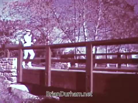 The still features a person in athletic attire jogging across a wooden bridge in a natural setting. The image, processed in black and white, captures the essence of physical activity and outdoor life, despite originally being in color. The contrast highlights the jogger and the bridge against the backdrop of trees.