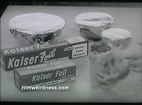 The still features various food containers covered with Kaiser Aluminum foil, prominently displaying the product packaging. The foil is shown in a quilted pattern, emphasizing its effectiveness for food preservation. Several containers, including bowls and jars, are arranged to showcase the foil's versatility and practicality in kitchen use. The visual conveys a sense of reliability and convenience associated with the product.