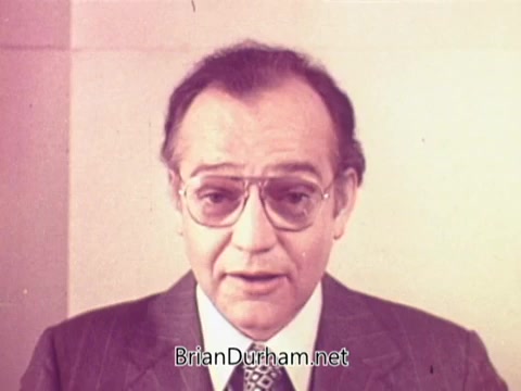 The still features a middle-aged man wearing glasses and a suit, addressing the camera. The background is simple and muted, directing focus to his face and message. The context of the image is related to a Bayer Aspirin commercial, suggesting a sales or health-related presentation. The overall tone appears informative and professional.