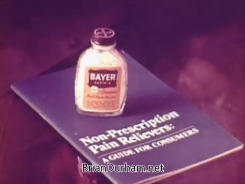 The still features a bottle of Bayer aspirin prominently displayed on top of a booklet titled 'Non-Prescription Pain Relievers: A Guide for Consumers.' The overall composition suggests a focus on consumer education regarding pain relief options. The color palette appears warm, emphasizing the branding of Bayer.