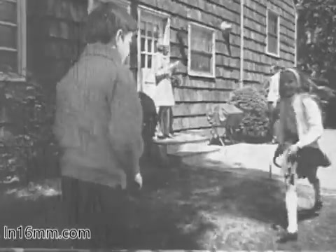The still from the 'Give Heart Fund PSA - 1960s' depicts two children outdoors, playing in front of a house. A girl, dressed in a skirt and holding a toy, is seen running towards a boy, who is turned towards her and appears to be engaged in play. In the background, a woman in a dress observes the scene from the porch, adding a sense of warmth and family life to the image. The setting reflects a typical suburban scene from the era, emphasizing innocence and camaraderie among children.
