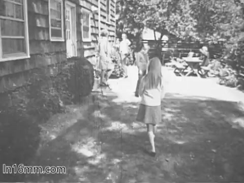 The still depicts a scene likely set in a backyard of a wooden house, capturing a moment from a PSA for the Give Heart Fund in the 1960s. In the foreground, a young girl in a light-colored sweater approaches a group of people in casual clothing. The background shows another group seated at a picnic table, suggesting a family or community gathering. The atmosphere appears warm and inviting, typical of public service announcements aimed at promoting heart health and community engagement during that era.