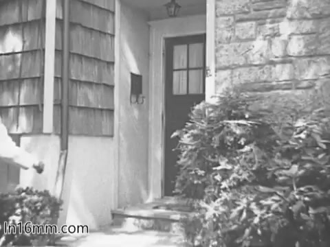 The still from the 1960s 'Give Heart Fund PSA' depicts a welcoming home entrance. Visible elements include a stone wall, wooden siding, and a front door with a window. Lush greenery surrounds the steps, contributing to a warm, inviting atmosphere typical of community-focused public service announcements from that era.
