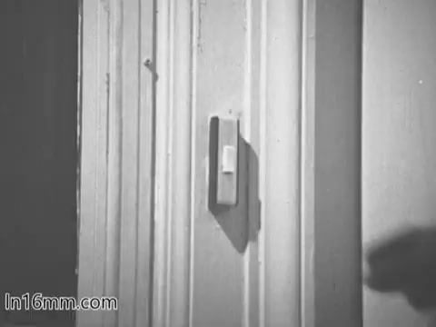 The still from the 'Give Heart Fund PSA - 1960's' depicts a hand reaching towards a wall-mounted doorbell. The image is monochromatic, highlighting the simplicity and directness of the scene, which likely sets the tone for a message about community and support related to heart health initiatives. The interaction suggests an invitation or call to action, typical of public service announcements of that era.