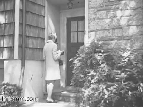 The still from the 1960s 'Give Heart Fund PSA' depicts a woman in a light-colored coat approaching a front door. She appears to be engaged in a charitable act, likely connected to the awareness campaign for heart health. The exterior suggests a suburban setting, with a mix of wood and stone on the house. Lush greenery frames the scene, creating a welcoming atmosphere.