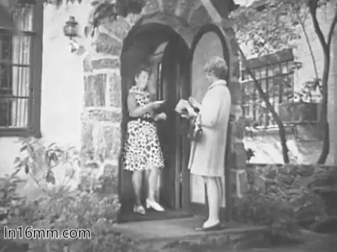 The still from the 'Give Heart Fund PSA - 1960s' depicts two women at the entrance of a house. One woman, dressed in a polka-dot dress, stands in the doorway, while the other, wearing a coat, is positioned outside the door. The setting suggests an informal conversation, possibly about the heart fund initiative, with a focus on community engagement. The scene is captured in black and white typical of the era's film style.