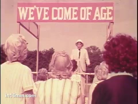 The still features a speaker in a light suit addressing a crowd from a stage adorned with a sign that reads 'WE'VE COME OF AGE.' The audience consists mostly of older women, seen from behind, suggesting a gathering or event focused on senior citizens, likely promoting empowerment or awareness within that demographic. The scene conveys a sense of community and celebration.