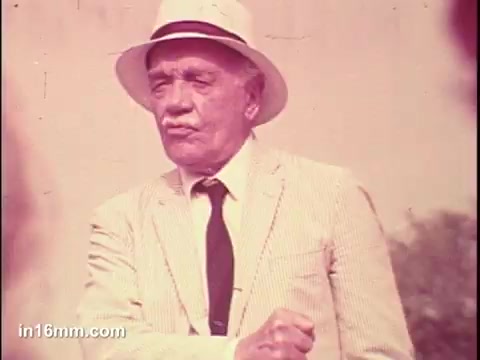 The still from the 1975 PSA for the National Council of Senior Citizens features an older man dressed in a light-colored striped suit and a white hat. He appears to be speaking or presenting, with a serious expression and a clenched fist, suggesting he is conveying an important message. The background is blurred, focusing attention on him and emphasizing his role in advocating for senior citizens. The colors have a vintage tone typical of 16mm film from that era.