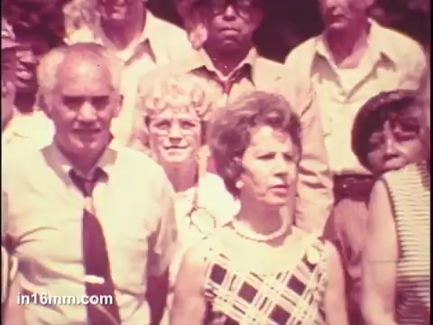 The still features a group of older adults, gathered outdoors, likely for a public service announcement related to senior citizens. Prominent figures include a man in a light-colored shirt and tie, and a woman in a plaid dress with pearl jewelry, indicating their roles as key speakers or representatives. The background shows a diverse assembly, suggesting community involvement and focus on senior citizen issues in 1975. The overall tone appears serious yet engaged, reflecting the era's emphasis on advocacy for the elderly.