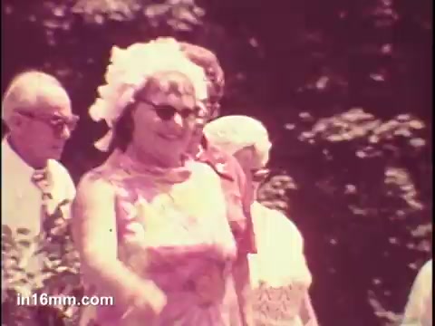 The still from the 1975 'National Council of Senior Citizens PSA' shows a cheerful group of seniors walking outdoors. The woman in the foreground, wearing sunglasses and a pink floral dress, appears to be enjoying herself. Other seniors are visible in the background, contributing to a vibrant and lively atmosphere, likely aimed at promoting positive aging and community among older adults.