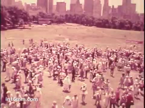 The still depicts a large gathering of senior citizens in a park setting, with a notable city skyline in the background. Many attendees are wearing hats, and the scene suggests a community event or rally aimed at promoting the interests of older adults. The atmosphere appears lively, reflecting a sense of camaraderie among the participants.