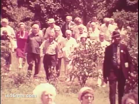 The still from the 'National Council of Senior Citizens PSA - 1975' features a large group of elderly individuals walking together outdoors. The scene captures a diverse gathering, showcasing seniors dressed in a variety of clothing styles typical of the 1970s. The setting appears to be a park or natural area, with greenery visible in the background. A few individuals are also dressed in formal attire, suggesting a community or organized event. The overall atmosphere reflects camaraderie and engagement among the senior citizens.