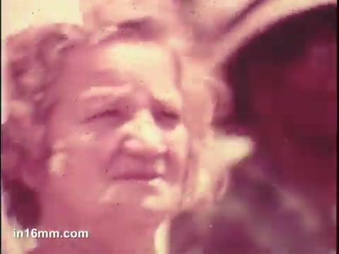 The still from the 'National Council of Senior Citizens PSA - 1975' features a close-up of an older woman with light-colored hair, looking intently at something off-camera. Her facial expression appears serious and contemplative. The background is somewhat blurry, suggesting a crowd or group of people, with a shadowy figure visible behind her. The overall tone of the image is warm but slightly faded, typical of vintage film quality.