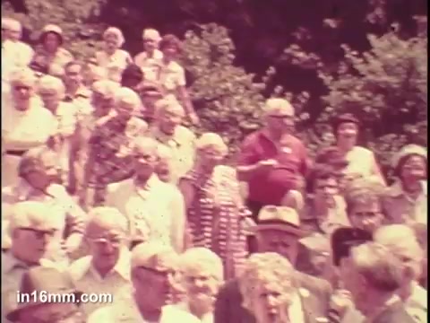 The still from the 1975 National Council of Senior Citizens PSA shows a large gathering of older adults, likely at an outdoor event. The crowd displays a variety of expressions and clothing typical of the era, capturing a sense of community among seniors. The background features greenery, indicating a lighthearted, social atmosphere. The image reflects themes of engagement and awareness concerning senior citizen issues during that time.