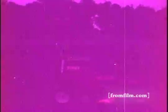 The still features a 16mm print from a 1970s Toro Riding Mower commercial. The image is characterized by a pinkish tint and possibly faded colors typical of older film stock. The mower appears to be in action, set against a blurred background of trees, capturing the outdoor setting commonly associated with lawn care. The overall look evokes a vintage aesthetic, reflecting the promotional style of that era.