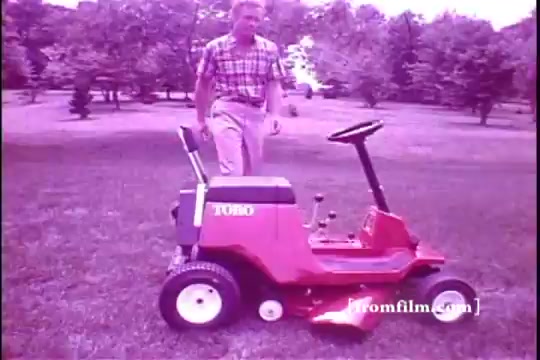 The still features a man in a plaid shirt standing next to a vibrant red Toro riding mower on a lush green lawn. The mower has a sleek, compact design typical of 1970s lawn equipment. The scene appears slightly washed out, indicative of the 16mm film quality, with a nostalgic ambiance reflecting that era. The background shows trees, enhancing the outdoor setting.