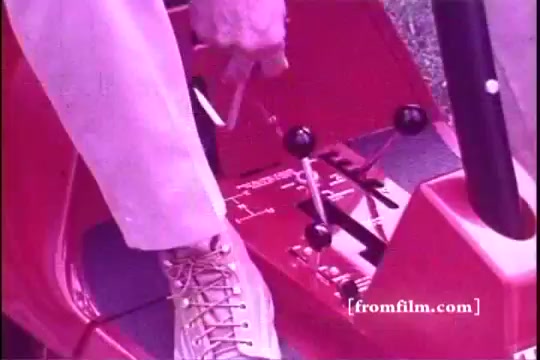 The still from the 16mm print features a close-up of a person's hand adjusting the controls on a Toro riding mower from the 1970s. The mower is a vibrant red, with visible levers and dials indicating its features. A beige shoe is shown, suggesting a casual attire typical of the era. The overall tone of the image has a vintage filter, enhancing the nostalgic feel of the commercial.