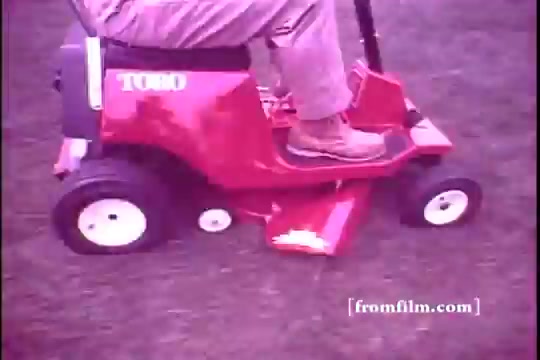 The still features a close-up of a person riding a red Toro riding mower in a grassy setting. The mower is compact, with a clear Toro logo on the side. The rider is wearing beige pants and brown shoes, indicating a casual outdoor appearance. The image captures a nostalgic 1970s aesthetic, characterized by soft focus and a slight color fade typical of 16mm film.