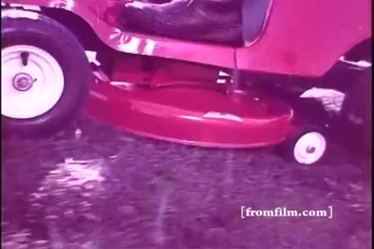 The still features a 16mm image of a Toro riding mower from the 1970s, showcasing its vibrant red color. Focused on the mower's mowing deck and wheel, the shot highlights the robust design and functionality, suggesting a scene of yard maintenance. The image quality reflects the vintage aesthetic typical of 16mm prints from that era.