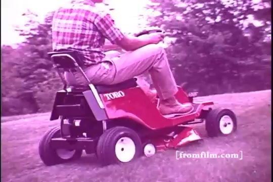 The still features a man operating a Toro riding mower, characteristic of the 1970s. He sits comfortably on the machine, which is red with a sleek design, and is depicted in a grassy outdoor setting. The image has a slightly faded quality typical of 16mm prints, capturing the nostalgic essence of the era. The man's plaid shirt and casual attire reflect the style of the time.