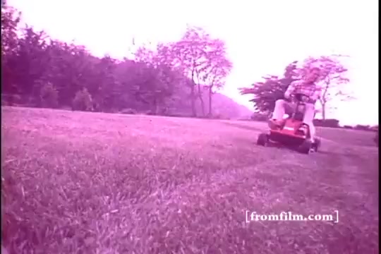 The still shows a mid-1970s Toro riding mower in action on a grassy lawn. A person is seated on the mower, navigating the terrain, with trees and hills in the background. The colors are soft and slightly faded, characteristic of a 16mm print, creating a nostalgic atmosphere. The focus is on the mower, highlighting its design and functionality.