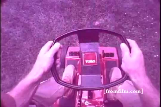 The still captures a top-down view of a Toro riding mower from the 1970s. Two hands are firmly gripping the steering wheel, and the mower's dashboard is visible, prominently featuring the Toro logo. The surrounding grass is neatly manicured, suggesting an active mowing scenario. The overall hue is slightly purple, indicative of the 16mm film's vintage qualities.