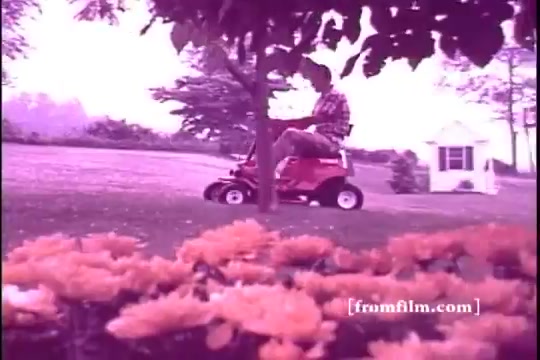 The still from the 16mm print of the 'Toro Riding Mower' commercial, likely from the 1970s, shows a man riding a bright red riding mower in a lush grassy area. In the foreground, vibrant orange flowers add color to the scene, while a quaint house can be seen in the background under a clear sky. The overall tone is nostalgic and reminiscent of suburban life during that era.