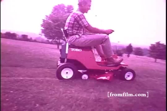 The still features a man riding a Toro riding mower across a grassy field. He is dressed casually in a plaid shirt and light pants, appearing cheerful as he operates the mower. The scene is likely from the 1970s, characterized by a vintage aesthetic with a slightly faded, pinkish hue typical of a 16mm film print. The background includes trees and an open landscape, suggesting a suburban or rural setting.
