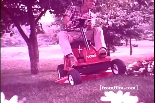 The still features a man riding a bright red Toro riding mower, typical of the 1970s aesthetic. He wears a plaid shirt, light-colored pants, and sandals, conveying a casual outdoor vibe. The mower is positioned on a grassy lawn, surrounded by trees and flowers, emphasizing a picturesque suburban scene. The film's color palette is warm, likely due to the vintage 16mm print, contributing to a nostalgic feel typical of promotional materials from that era.