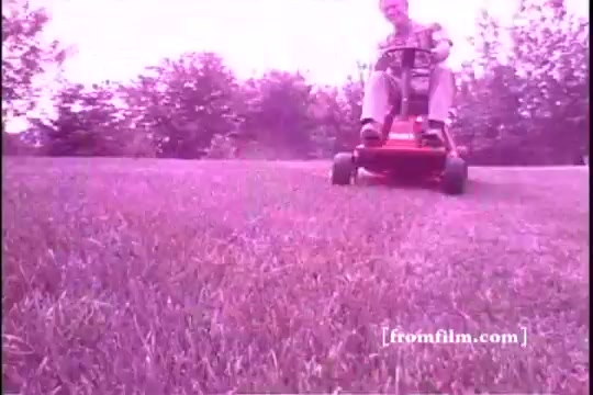 The still features a man operating a Toro riding mower across a grassy lawn. The image has a vintage aesthetic, typical of 16mm film from the 1970s, characterized by its slightly muted colors and a soft focus. The scene captures a moment of leisure and functionality, suggesting the mower's effectiveness in maintaining a well-kept yard. In the background, trees can be seen, hinting at a suburban setting.