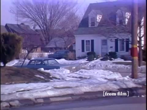 The still captures a winter suburban street scene featuring two men near a parked car, with a yellow fire hydrant visible alongside a snow-covered landscape. The camera angle is positioned from the street, offering a view of a home in the background, framed by barren trees.