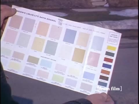 The film still depicts a wintertime suburban street where two men review a color palette held aloft, with a glimpse of a typical home and a yellow fire hydrant in the background. The camera angle appears to focus on the palette while capturing the suburban setting subtly out of focus.