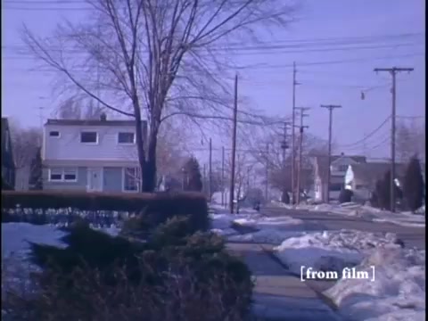 The still depicts a winter suburban scene with a residential house in the background, flanked by telephone poles and a snow-covered sidewalk. In the foreground, a neatly trimmed hedge leads the viewer's eye across the frame, capturing the tranquil atmosphere of the 1960s suburban setting.